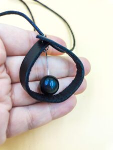 Hand holding a handmade black leather pendant necklace with a glossy black bead suspended inside a teardrop-shaped leather loop on a black cord.