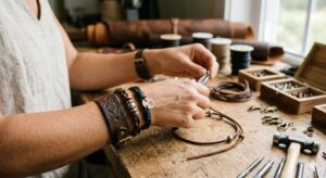 Close-up of an artisan's hands using pliers to craft DIY leather bracelets at a rustic wooden workbench filled with tools, spools of cord, and beads.