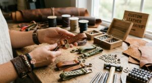 An artisan's hands holding a custom brown leather keychain stamped with the word "ADVENTURE" over a rustic wooden workbench. The table is filled with finished leather loop and braided keychains, metal letter stamps, a hammer, and raw leather scraps.