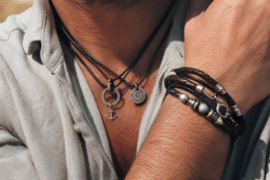 Close-up of a person wearing layered leather cord necklaces with metallic charms and stacked leather and bead bracelets on the wrist, captured in warm natural outdoor lighting with a soft blurred background.